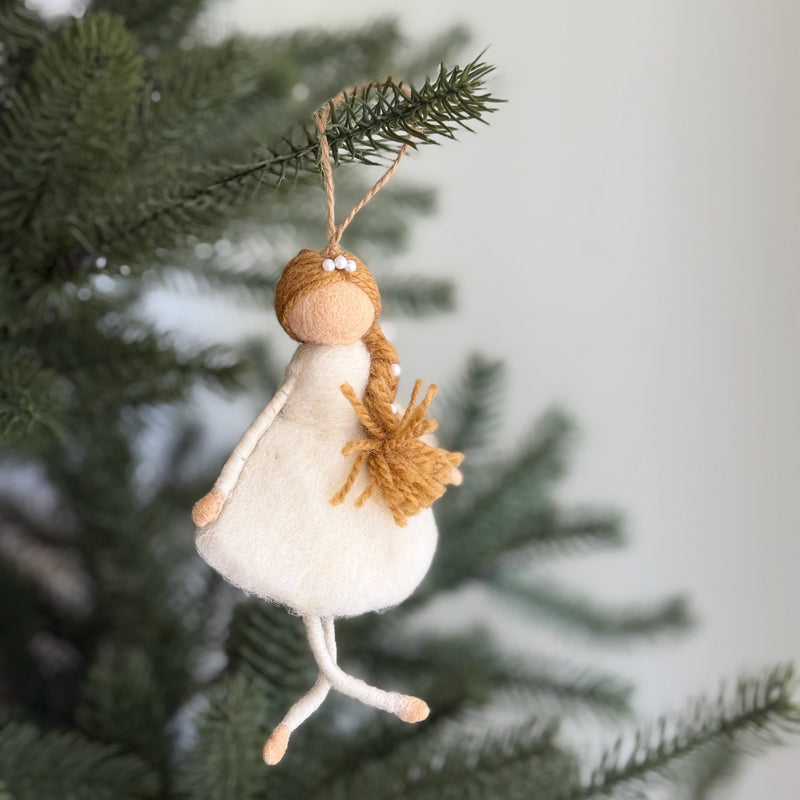 A felt ballerina ornament hangs gracefully from a green Christmas tree branch. This charming handmade felt ornament features a round, light beige head with white beads and braided brown yarn. Her off-white wool felt dress and beige felt legs complete the festive design, ready to add whimsy to your holiday decor.