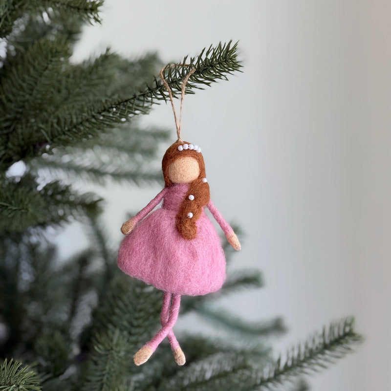 A delicate felt ballerina ornament is suspended by a natural jute string on a Christmas tree, set against a soft, light backdrop. This hand-felted wool ballerina features a soft pink wool felt dress, light peach upper body and legs. A brown felted braid adorned with small white beads completes the charming and whimsical design.