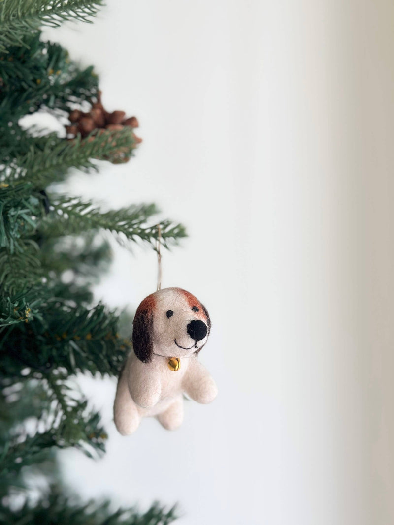 A charming felt beagle Christmas ornament with a tiny golden bell hangs gracefully from a green Christmas tree adorned with pine cones. This handcrafted felt beagle features a predominantly white body with brown and dark brown patches on its head and ears. The soft texture and jute string for hanging add a joyful touch to holiday decor.