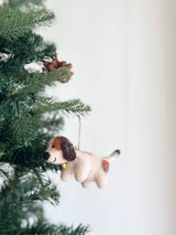 A charming felt beagle ornament, needle felted and adorned with a gold bell, hangs on a lush Christmas tree, nestled among green needles. This festive beagle features a cream-colored body with brown markings, a black nose, and a jute string for hanging.