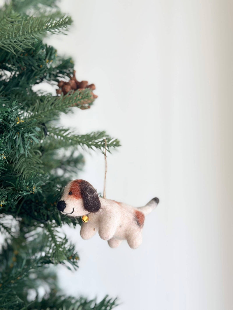 A charming felt beagle ornament, needle felted and adorned with a gold bell, hangs on a lush Christmas tree, nestled among green needles. This festive beagle features a cream-colored body with brown markings, a black nose, and a jute string for hanging.