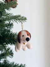 A needle felted beagle ornament, crafted from soft wool felt, hangs on a Christmas tree with green needles, set against a minimalist white backdrop. This charming beagle features a cream-colored body, brown head markings, and floppy brown ears. A golden bell is around its neck, with a jute string attached on top for hanging.