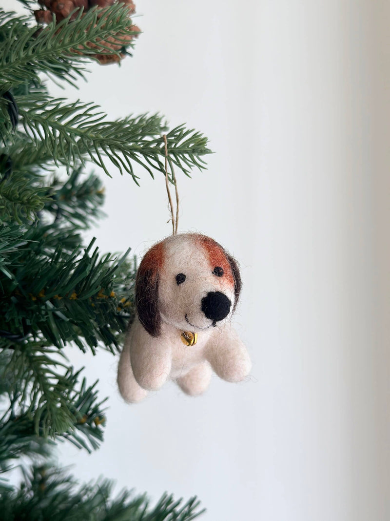 A needle felted beagle ornament, crafted from soft wool felt, hangs on a Christmas tree with green needles, set against a minimalist white backdrop. This charming beagle features a cream-colored body, brown head markings, and floppy brown ears. A golden bell is around its neck, with a jute string attached on top for hanging.