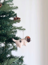 A charming felt beagle ornament, adorned with a small golden bell, hangs on a festive Christmas tree. The unique beagle ornament has a fluffy, off-white body, a distinctive black spot, and a brown patch on its head, with a delicate cotton string for hanging. Pine needles and brown pine cones surround it, evoking holiday warmth and joy.