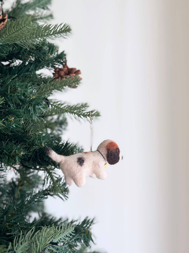 A charming felt beagle ornament, adorned with a small golden bell, hangs on a festive Christmas tree. The unique beagle ornament has a fluffy, off-white body, a distinctive black spot, and a brown patch on its head, with a delicate cotton string for hanging. Pine needles and brown pine cones surround it, evoking holiday warmth and joy.