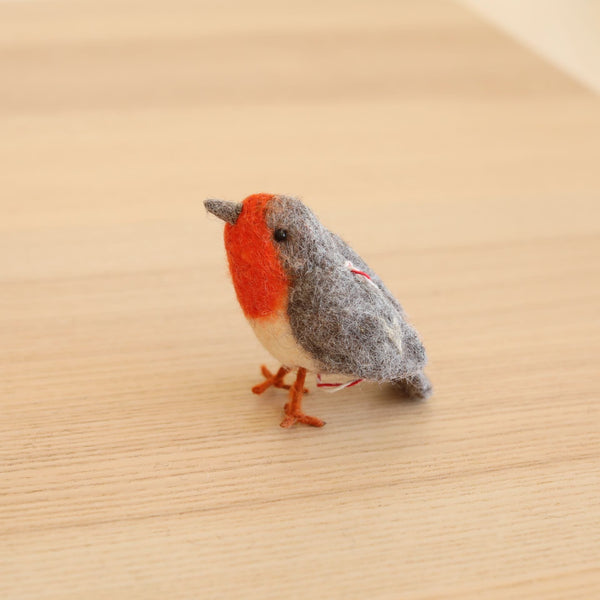 A handmade wool felt robin ornament is prominently displayed on a neutral surface. This charming 3D bird features the distinct colors of a robin, including a grey head, brown beak, bright red chest, and brown feet. Individually needle felted and hand-stitched, it has subtle white and red stitching, perfect for holiday decor.