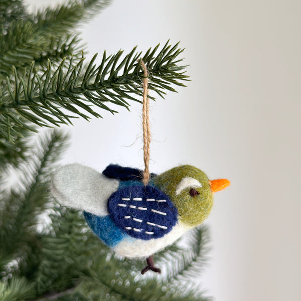 A charming handmade felt Blue Bird ornament hangs on a Christmas tree branch against a simple white background. This unique Christmas ornament features a delightful bird with a green head, a white and blue body, and a dark blue wing detailed with white stitches. An orange beak and a natural jute string complete this whimsical design, ready to add cheer to holiday decor.