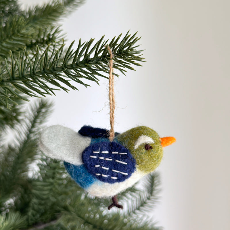 A charming handmade felt Blue Bird ornament hangs on a Christmas tree branch against a simple white background. This unique Christmas ornament features a delightful bird with a green head, a white and blue body, and a dark blue wing detailed with white stitches. An orange beak and a natural jute string complete this whimsical design, ready to add cheer to holiday decor.