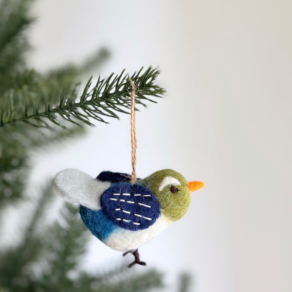 A blue bird felt ornament is gracefully suspended from a vibrant green Christmas tree branch. This charming handmade felt ornament features a green head with a white eye accent, a pointed orange beak, and a fluffy white wing. Its body transitions from dark to light blue, accented by a navy blue patch with white stitches on its back, leading to delicate brown felt feet and a jute hanging string.