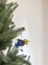 A handcrafted felt blue bird ornament hangs from a pine branch, against a Christmas tree and a white wall. The soft felt ornament features a round body with a white belly, a blue and white patterned wing, an olive-green head, and an orange beak. It includes a jute string for easy hanging.