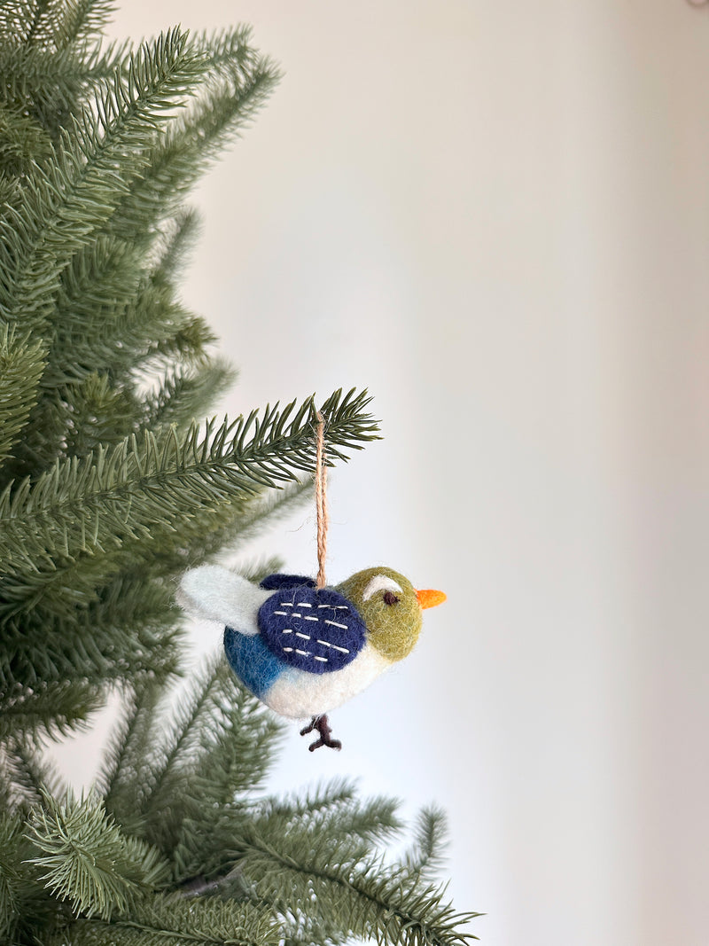 A handcrafted felt blue bird ornament hangs from a pine branch, against a Christmas tree and a white wall. The soft felt ornament features a round body with a white belly, a blue and white patterned wing, an olive-green head, and an orange beak. It includes a jute string for easy hanging.