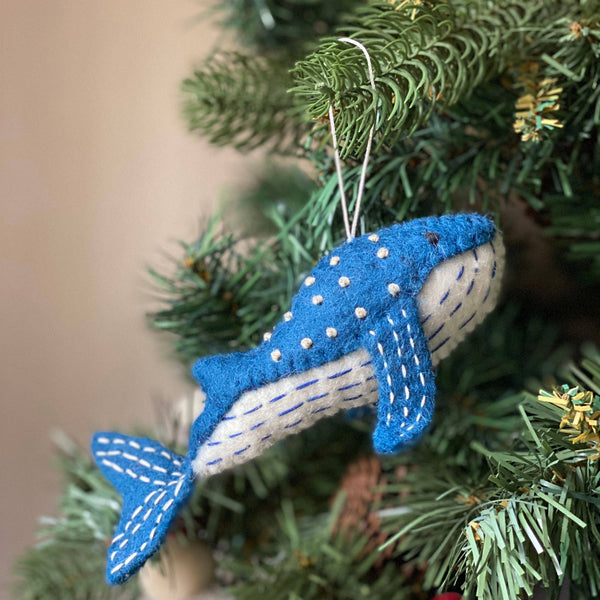 A needle felted blue whale felt ornament hangs from the green branches of a Christmas tree. The vibrant blue whale features small, raised white dots on its back and white stitched details on its fins and lower body, with a white string for hanging.