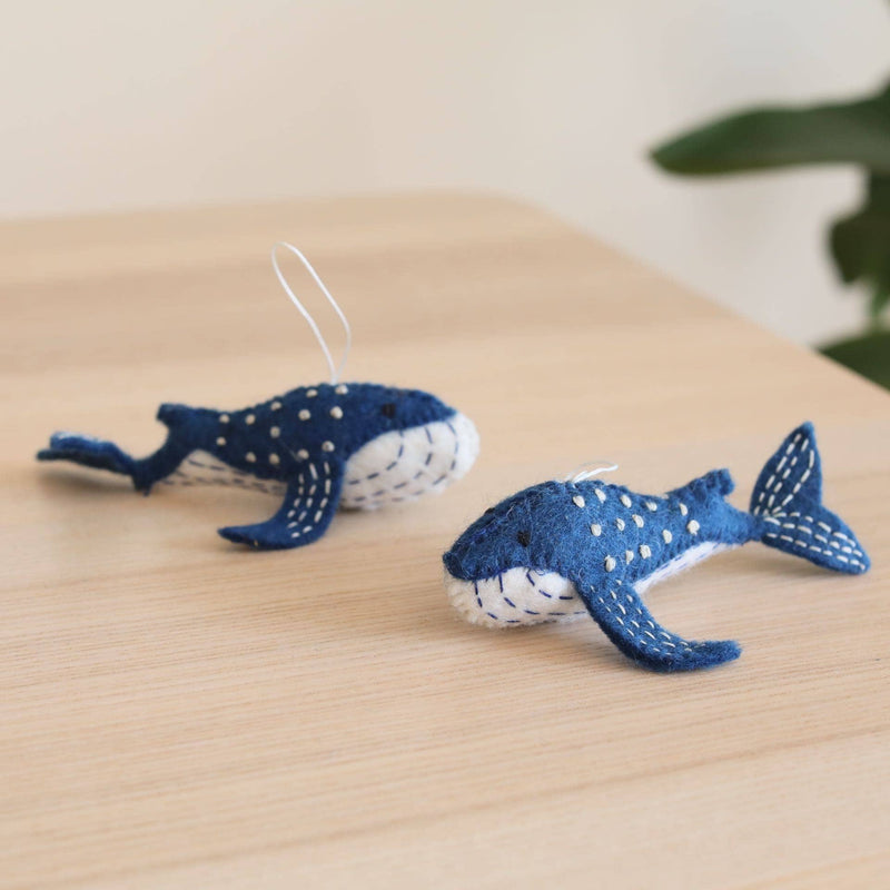 Two needle felted Blue Minke Whale ornaments rest on a light wood surface, set against a soft, neutral background with a hint of greenery. Crafted from blue organic wool, each whale features distinctive shapes with flippers, a tail, and a white underbelly, all highlighted by contrasting white stitching. A white cotton string is attached, ready for hanging, showcasing their detailed craftsmanship as unique Christmas tree ornaments.