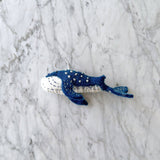A handmade, needle felted Blue Minke Whale ornament rests on a mottled white and gray marble surface. This organic wool whale ornament is predominantly blue with white accents on its underside. The blue features white embroidered spots, while the white underside has blue stitches, with a white cotton string for hanging.