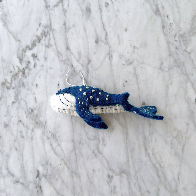 A handmade, needle felted Blue Minke Whale ornament rests on a mottled white and gray marble surface. This organic wool whale ornament is predominantly blue with white accents on its underside. The blue features white embroidered spots, while the white underside has blue stitches, with a white cotton string for hanging.