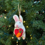 A charming white wool felt bunny Christmas ornament, holding a vibrant red heart embellished with a small felt carrot, hangs gracefully on a lush Christmas tree adorned with delicate lights. This intricately hand-stitched bunny features pink ear accents, a pink nose, and embroidered black whiskers. A jute string is attached for hanging.