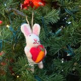A charming needle felted wool bunny ornament, holding a vibrant red heart, hangs on a festive Christmas tree. The soft white bunny features long ears with pink interiors and an embroidered face with a red mouth and black whiskers. It is delicately placed among green tree needles and glowing fairy lights, with other ornaments visible in the cozy background.