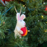 A cute felt bunny holding a heart Christmas tree ornament hangs on a festive tree with green needles and subtle lights. This handcrafted felt bunny ornament is white with fluffy texture, pink inner ears, and black eyes. It clasps a vibrant red and orange wool felt heart, perfect for holiday decor.