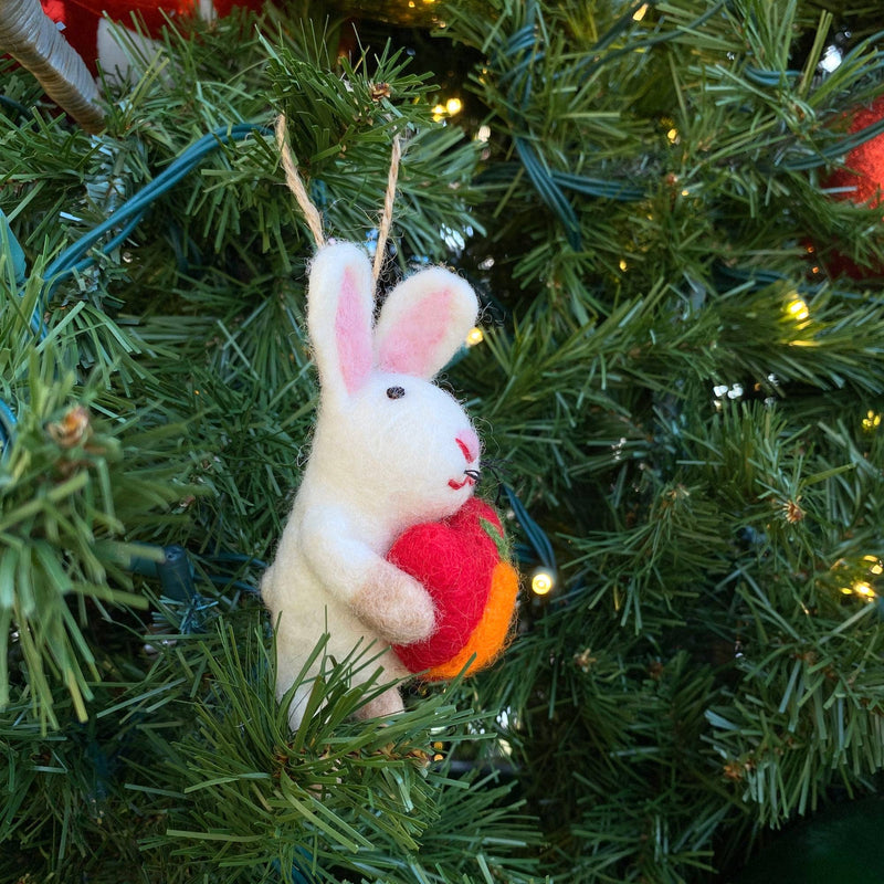 A cute felt bunny holding a heart Christmas tree ornament hangs on a festive tree with green needles and subtle lights. This handcrafted felt bunny ornament is white with fluffy texture, pink inner ears, and black eyes. It clasps a vibrant red and orange wool felt heart, perfect for holiday decor.