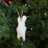 A handmade felt bunny ornament, holding a heart, hangs on a green Christmas tree adorned with warm lights. The white, needle-felted bunny is depicted from the back, showcasing its long ears, a small fluffy tail, and tan accents on its feet. A jute string on its head allows it to hang charmingly from the branch.