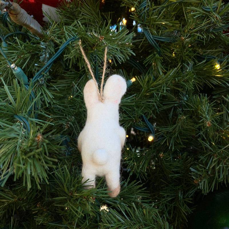 A handmade felt bunny ornament, holding a heart, hangs on a green Christmas tree adorned with warm lights. The white, needle-felted bunny is depicted from the back, showcasing its long ears, a small fluffy tail, and tan accents on its feet. A jute string on its head allows it to hang charmingly from the branch.