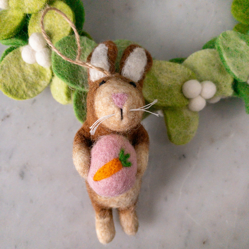 A handcrafted wool felt bunny Christmas ornament is displayed against a light gray backdrop, partially framed by a green mistletoe wreath. This whimsical brown and beige bunny holds a pink heart with a green leaf, features a pink nose and white whiskers, and has a jute string attached for easy hanging.