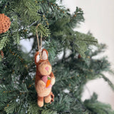 A wool felt bunny Christmas ornament holding a heart is displayed on a green Christmas tree. The charming bunny features light and dark brown hues, long ears, a pink nose, and a sweet expression. It holds a pink heart with a small orange carrot in its paws and hangs by a jute string.