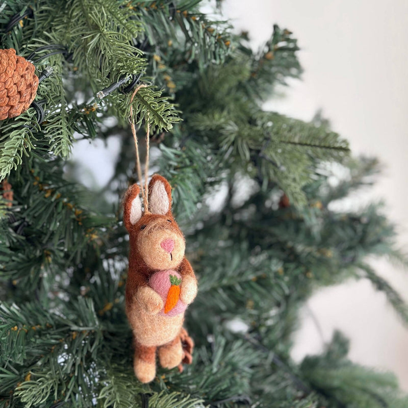 A wool felt bunny Christmas ornament holding a heart is displayed on a green Christmas tree. The charming bunny features light and dark brown hues, long ears, a pink nose, and a sweet expression. It holds a pink heart with a small orange carrot in its paws and hangs by a jute string.