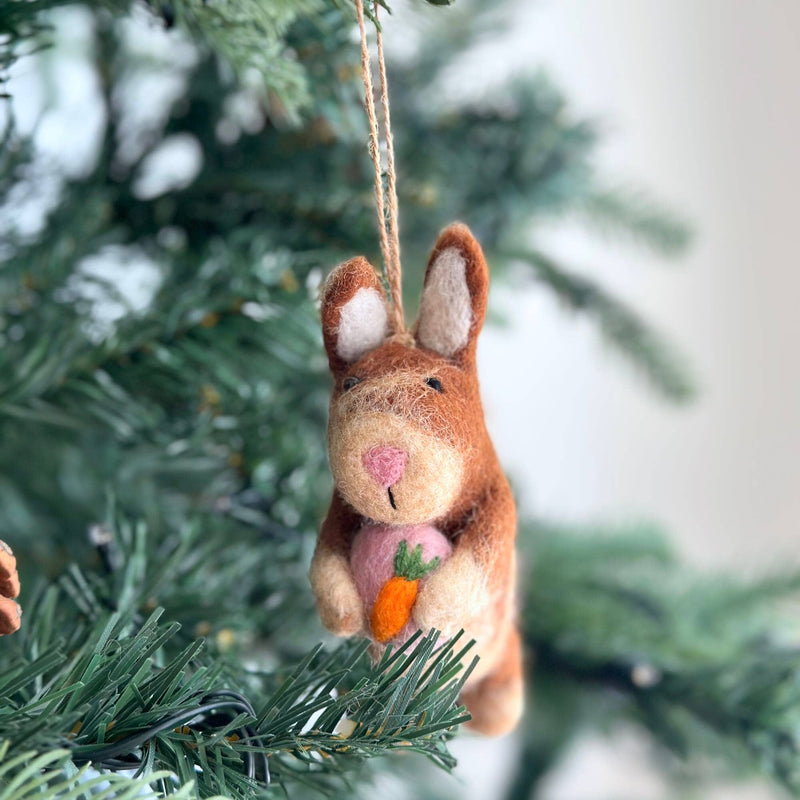 A felt bunny ornament, holding a pink heart, hangs gracefully on a lush green Christmas tree. The brown bunny features lighter beige accents on its face and inner ears. A jute string from its head allows this charming Christmas ornament to be nestled among the deep green needles, evoking a cozy, festive atmosphere.