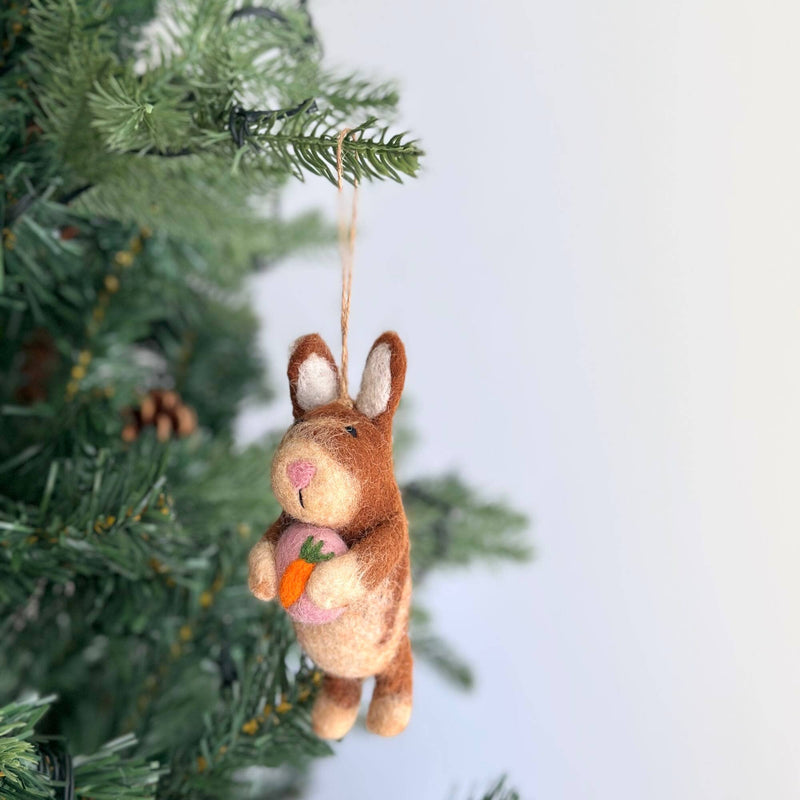 A handmade felt bunny ornament, holding a heart, hangs charmingly from a Christmas tree branch adorned with green needles and pine cones. Crafted from organic wool, the bunny is predominantly brown with lighter accents on its ears, paws, and belly, and features a pink nose. It sweetly holds a pink heart, adding a whimsical touch to the festive holiday decor.