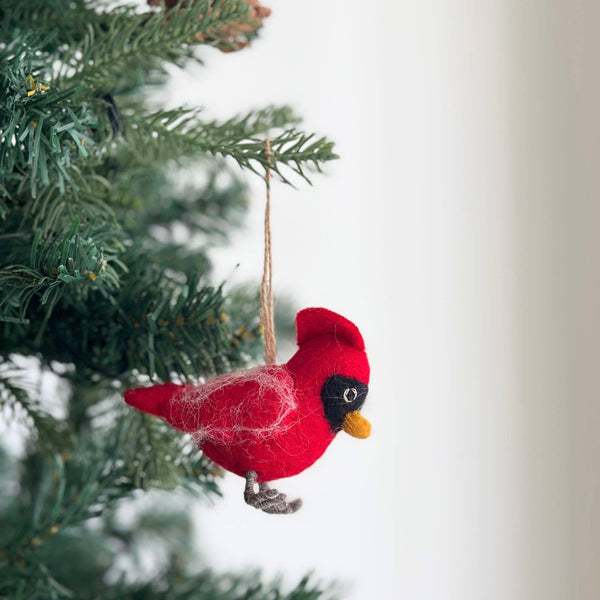 A red felt cardinal ornament hangs on a vibrant green Christmas tree, set against a bright, airy backdrop. This handcrafted felt cardinal features a red body with a yellow beak and intricate gray felt feet. Subtle white felt accents and a black felt accent around the eye enhance its textured body. A jute string allows for easy hanging.