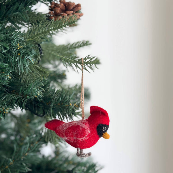 A vibrant red felt cardinal ornament with a yellow beak, black eye markings, and gray feet hangs from a green Christmas tree branch by a rustic jute string. Decorative white accents dot its body. Above the handmade felt cardinal ornament rests a brown pine cone, all set against a bright white background, creating a festive holiday display.