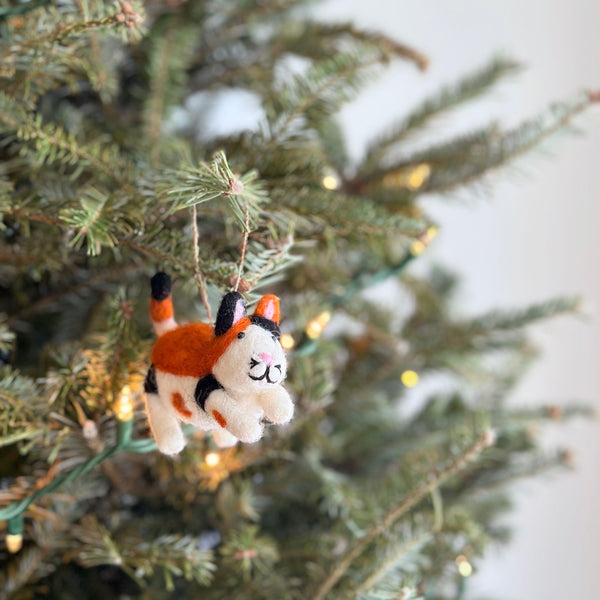 A needle felted calico cat Christmas ornament hangs on a Christmas tree, creating a warm, festive ambiance. This 3D felt ornament features a distinct white, orange, and black calico pattern, perky ears, and stitched facial details. Crafted from 100% New Zealand sheep wool, it dangles from a jute string among the tree's green needles and soft fairy lights.