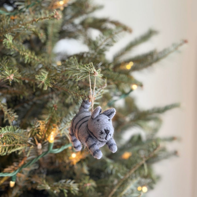 A charming wool felt gray tabby cat ornament hangs on a lit Christmas tree, evoking a warm, festive holiday mood indoors. This handcrafted needle felted Christmas ornament features a 3D design with gray felt, darker gray stripes, and perky ears, ready to adorn your tree.