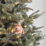A needle felted gray tabby cat ornament hangs on a Christmas tree. This 3D wool felt Christmas ornament features a textured gray body with darker tabby stripes, suspended by a jute string. Green tree needles and soft, glowing string lights provide a festive backdrop, adding a cozy touch to holiday decor.