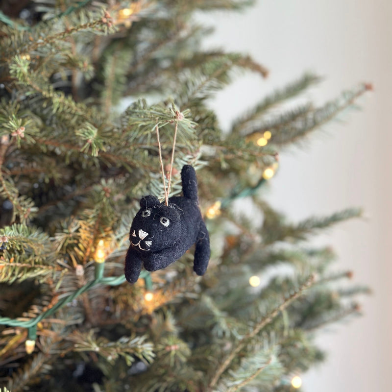 A charming needle-felted black cat Christmas ornament hangs on a Christmas tree, illuminated by warm white lights. This 3D-designed felt ornament is crafted from 100% New Zealand sheep wool and features delicate white thread whiskers and eyes. It is suspended from a natural jute string, adding to its cozy appeal.