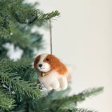 A charming felt Cavalier King Charles Spaniel dog ornament is displayed on a Christmas tree illuminated by warm, festive lighting. This unique handmade felt ornament features a white body, brown head markings, a golden bell around its neck, and a jute string for hanging.