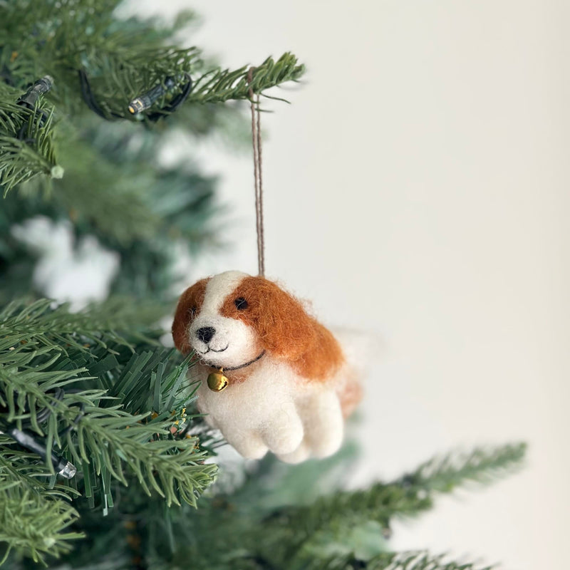 A charming felt Cavalier King Charles Spaniel dog ornament is displayed on a Christmas tree illuminated by warm, festive lighting. This unique handmade felt ornament features a white body, brown head markings, a golden bell around its neck, and a jute string for hanging.