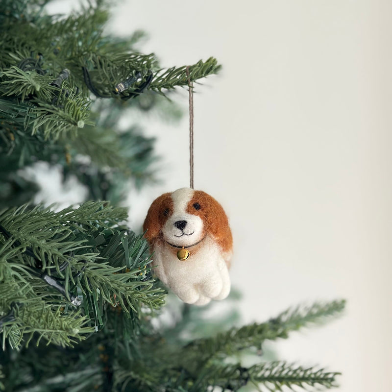 A handcrafted felt Cavalier King Charles Spaniel dog Christmas ornament hangs from a subtly lit Christmas tree branch. This brown and white felt ornament is meticulously shaped with distinctive markings and features. A delicate golden bell hangs from a black string around its neck, adding a festive touch to the warm seasonal scene, suspended by a jute string.