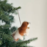 A handmade felt Cavalier King Charles Spaniel dog ornament hangs on a green Christmas tree with small white lights, evoking warmth and holiday cheer. The white and brown dog ornament features a small golden bell around its neck and is suspended by a jute string.