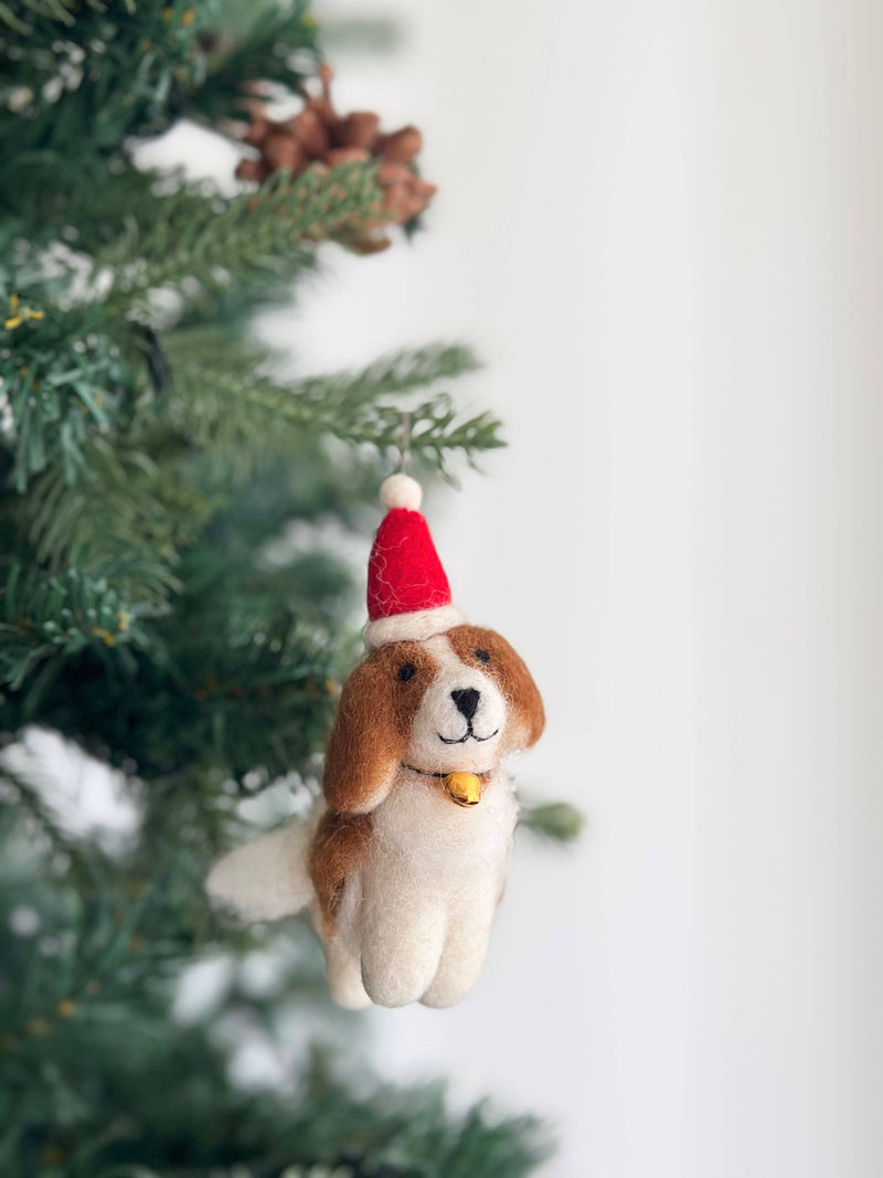 A handcrafted white and light brown wool felt Cavalier King Charles Spaniel dog ornament hangs charmingly on a lush, festive Christmas tree. This unique Christmas ornament wears a tiny red Santa hat with a white pompom and trim, and a small gold bell on its collar, adding a playful holiday touch.