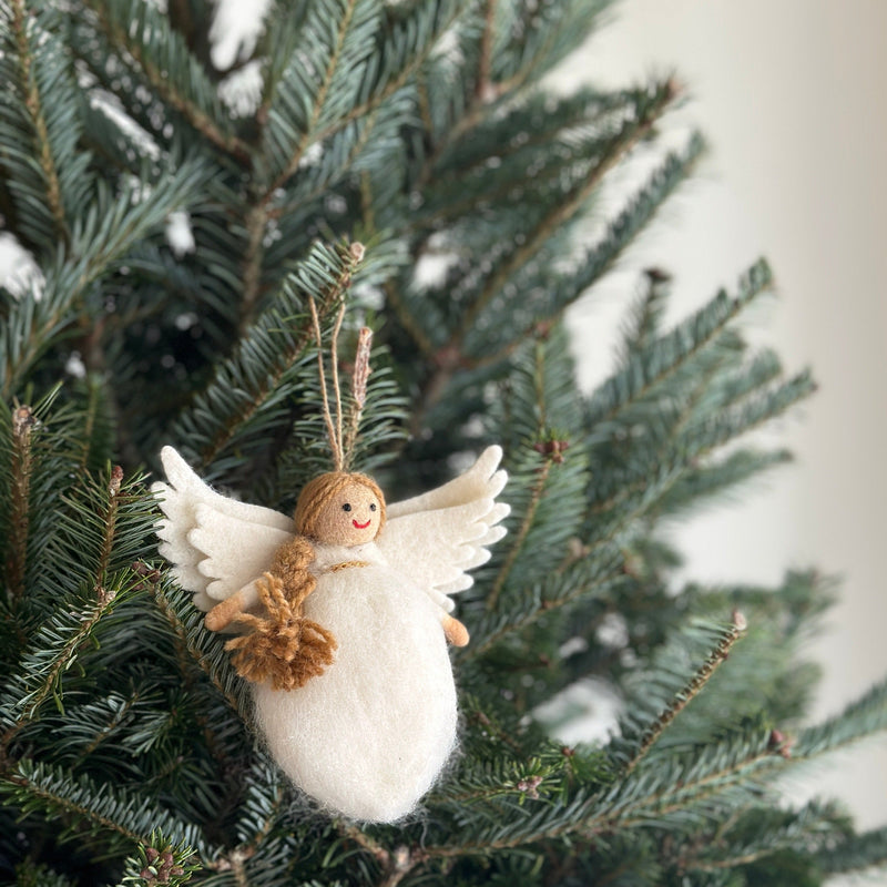 A handmade needle felted Christmas angel ornament hangs on a lush green Christmas tree. The charming angel features delicate white wings, a round white body, brown hair, a soft smile, and rosy cheeks, with a jute string attached for easy hanging.