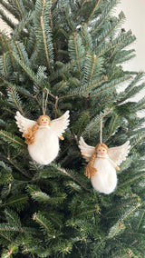 Two wool felt Christmas angel ornaments hang on the green branches of a Christmas tree. Each white angel ornament features a plump body, layered wings, and a head adorned with a golden wool braid. A jute string is attached for hanging these unique Christmas ornaments, creating a festive display.