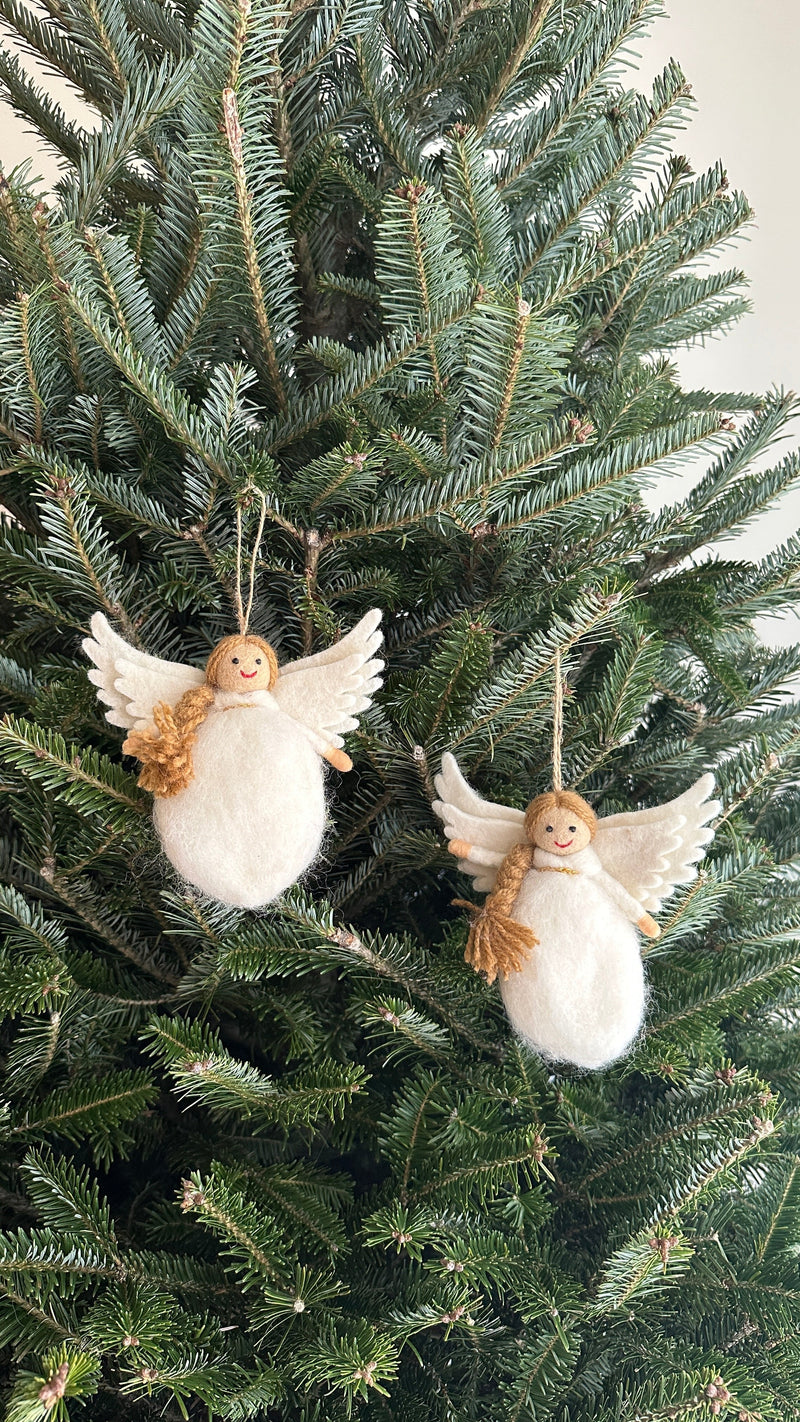 Two wool felt Christmas angel ornaments hang on the green branches of a Christmas tree. Each white angel ornament features a plump body, layered wings, and a head adorned with a golden wool braid. A jute string is attached for hanging these unique Christmas ornaments, creating a festive display.