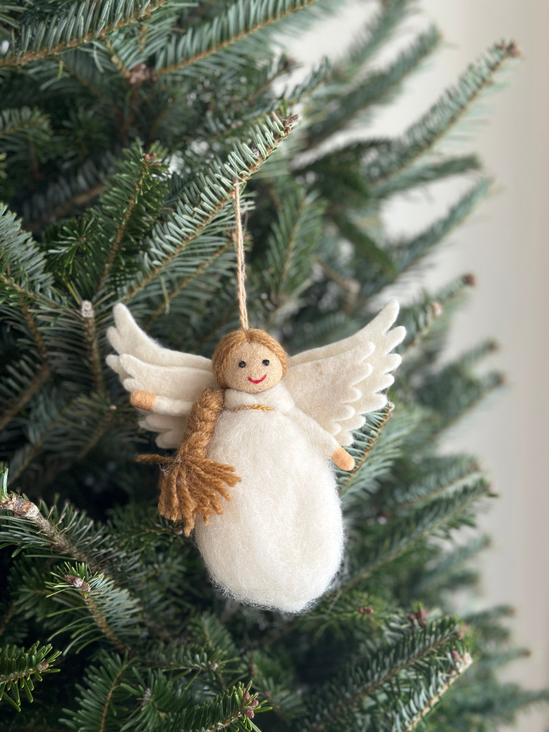 A charming wool felt angel ornament hangs gracefully from a Christmas tree branch. The unique Christmas ornament features delicate white wool felt wings and body, complemented by flaxen hair, dot eyes, and a stitched smile. A jute string is attached for easy hanging.