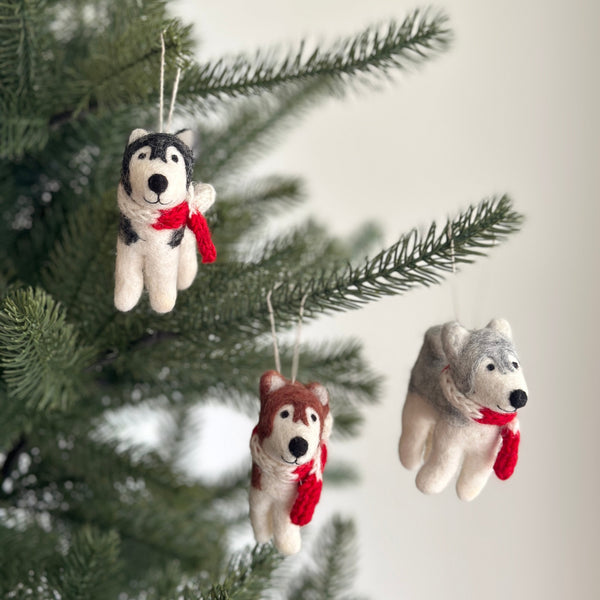 Three Christmas husky felt ornaments with red scarves are charmingly displayed on a vibrant green Christmas tree. Each handcrafted felt ornament hangs from a white cotton string. One husky is white with black markings, another white with brown, and the third white with grey. Each adorable husky wears a bright red scarf, adding a festive pop of color.