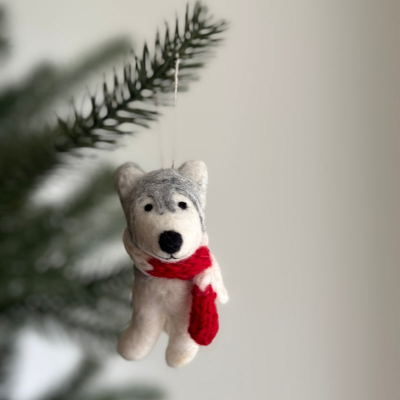 A handcrafted felt husky Christmas ornament with a red scarf hangs on a blurred dark green Christmas tree. This unique husky felt ornament features detailed gray and white textured fur, upright ears, and a black nose. A white cotton string is attached for hanging, creating a calm and festive ambiance.