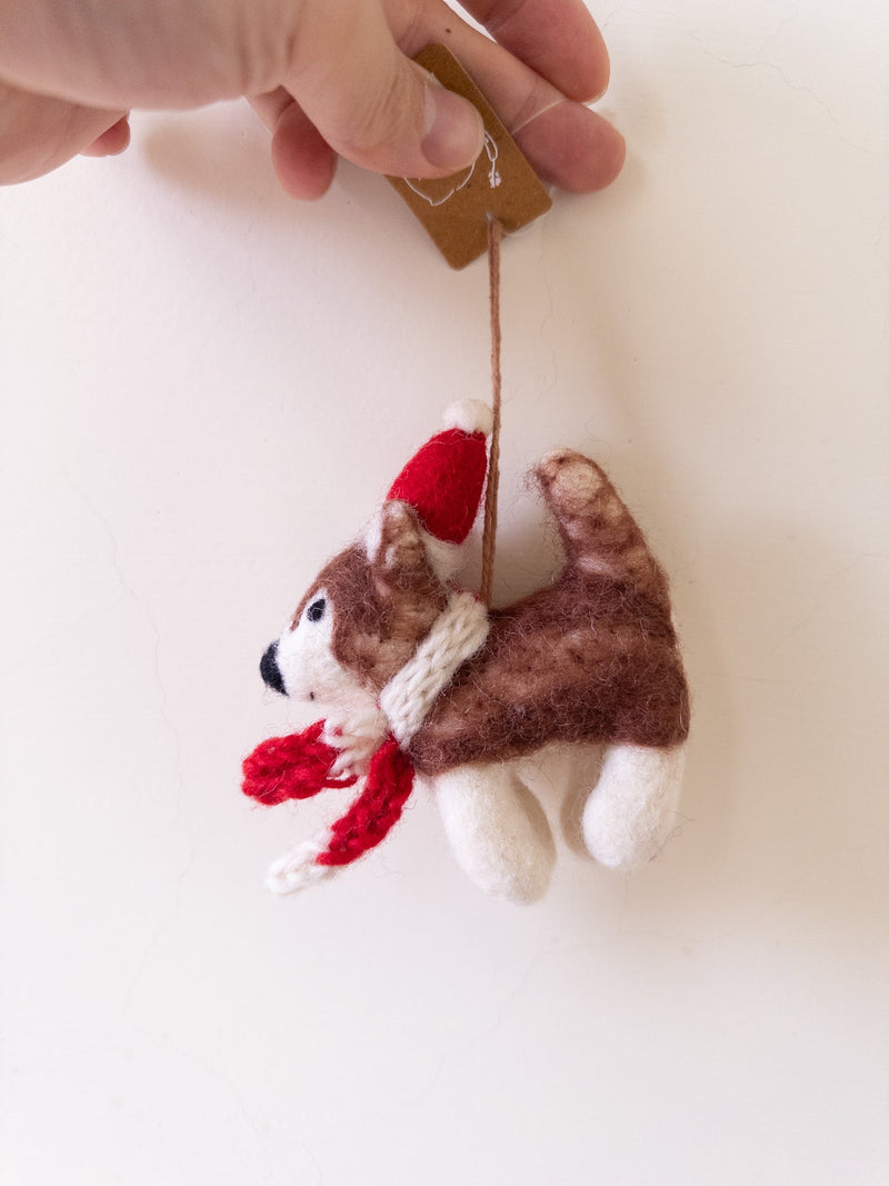 A handmade felt Christmas husky ornament is held by a female hand, suspended against a plain white background. This charming felt husky features a brown and white body with a black nose, wearing a festive red Santa hat and a matching scarf around its neck.