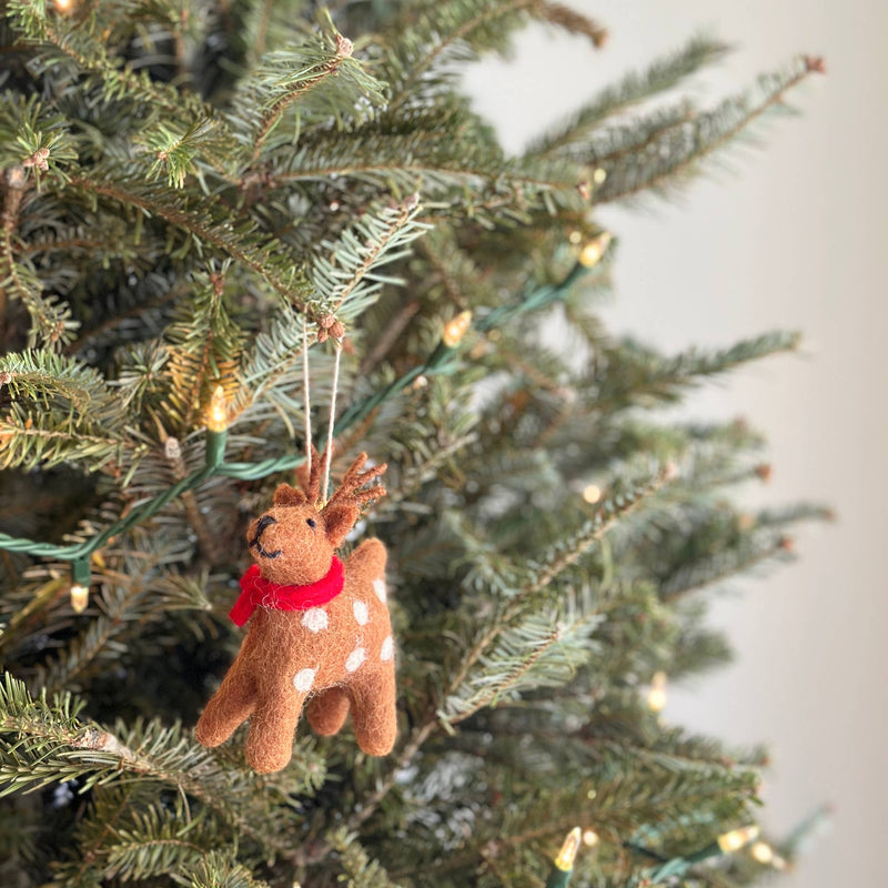 A handmade wool felt reindeer ornament hangs on a Christmas tree, nestled among green needles and festive lights. The light brown reindeer ornament features small white dots, a tiny red scarf, and delicate antlers, with a white cotton string for hanging.