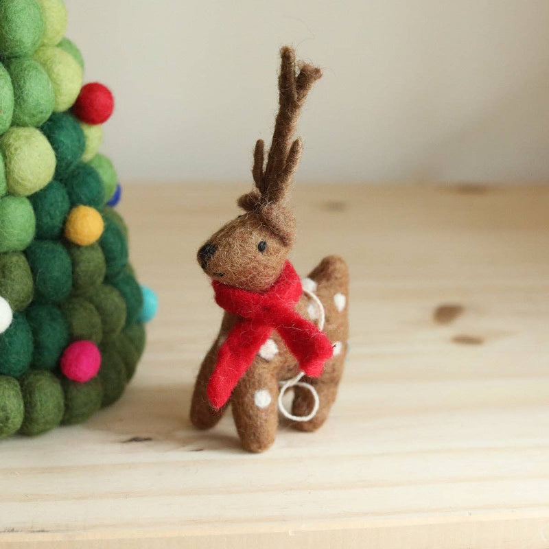 A handcrafted felt reindeer Christmas ornament rests on a light wooden surface, positioned next to a small green felt Christmas tree, evoking a festive mood. The brown wool felt reindeer has white spots and is adorned with a bright red scarf. Its textured, needle-felted construction is evident, with a white cotton string attached for hanging.